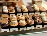 A display of various cupcakes neatly arranged on a wooden tray, each with different toppings and labels. The cupcakes include flavors like S'mores, Butterscotch Honeycomb, Salted Caramel, Milk Chocolate Banana, Chocolate Hazelnut, and Earl Grey. The toppings range from cookies and cream to nuts and swirls of frosting.
