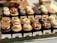 A display of various cupcakes neatly arranged on a wooden tray, each with different toppings and labels. The cupcakes include flavors like S'mores, Butterscotch Honeycomb, Salted Caramel, Milk Chocolate Banana, Chocolate Hazelnut, and Earl Grey. The toppings range from cookies and cream to nuts and swirls of frosting.