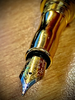 A close-up view of a shiny, golden fountain pen nib resting on a wooden surface. The nib features intricate engravings, and the metallic finish reflects light with a glossy appearance.