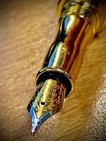 A close-up view of a shiny, golden fountain pen nib resting on a wooden surface. The nib features intricate engravings, and the metallic finish reflects light with a glossy appearance.
