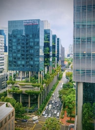 Tall modern buildings with glass facades and lush greenery integrated into their structure line a wide street with traffic below. Trees and plants adorn the buildings, creating a blend of urban and natural elements. The scene is vibrant and dynamic, with a mix of nature and architecture.
