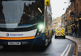 A double-decker bus with a visible Stagecoach logo is driving down a wet city street, reflecting the overcast sky. Behind the bus, a white van with red and yellow chevron markings is visible. There are buildings lining the narrow street, with one person walking on the sidewalk.