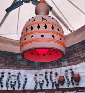A handcrafted terracotta lamp with intricate cut-out designs hangs from the ceiling. The lamp emits a warm pinkish-red glow, creating a soft ambiance. In the background, a brick wall supports various hanging decorative items, adding a rustic charm.