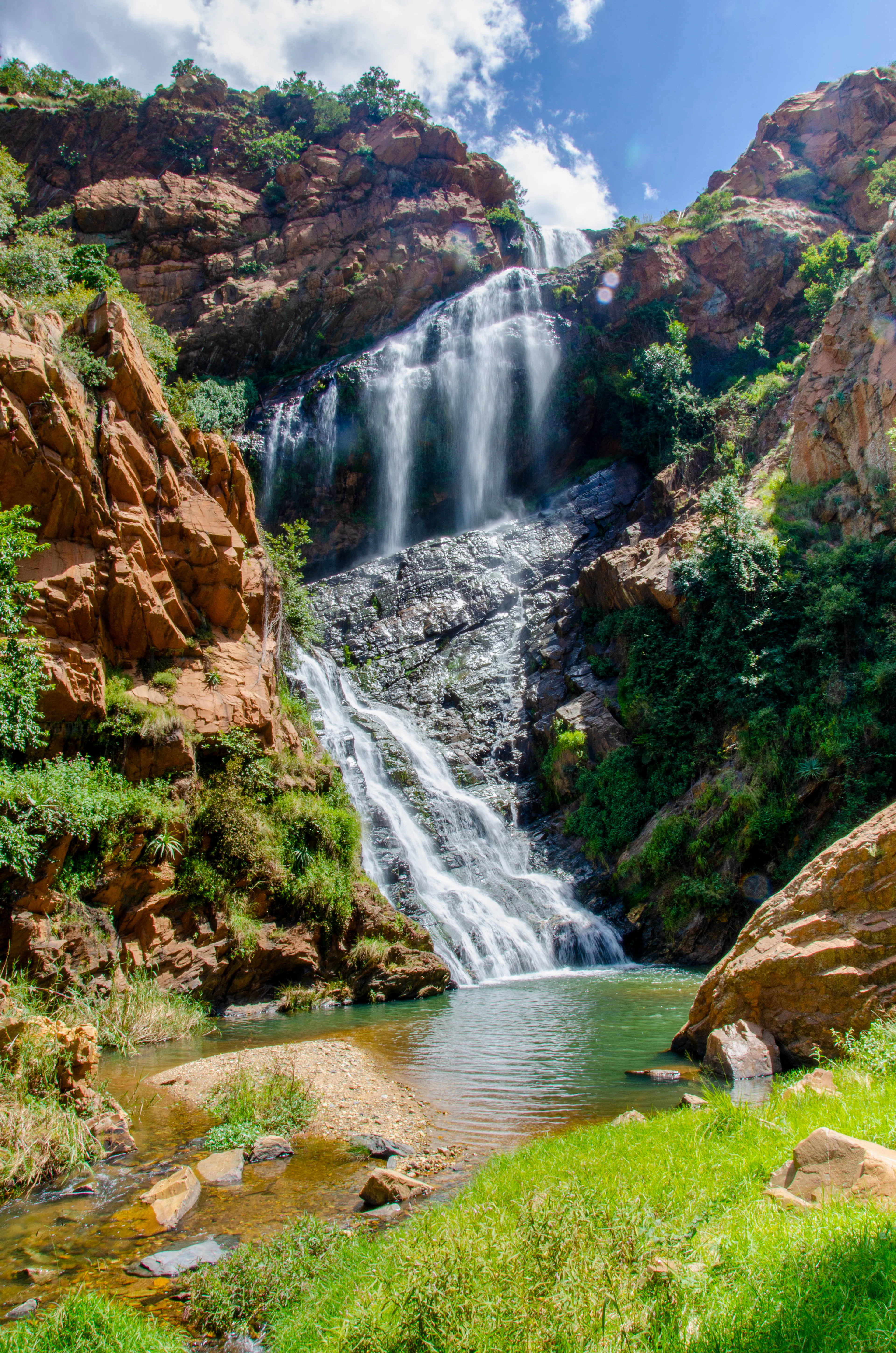 The waterfall at Walter Sisulu National Botanical Garden on Johannesburg's western edge, dropping in two silver tiers down a gorge of red quartzite cliffs into a green plunge pool, framed by bright grass and sunlit blue sky.