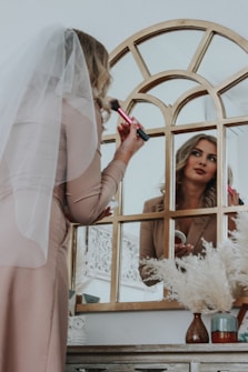 A woman is applying makeup in front of a decorative mirror. She is wearing a light-colored dress with a veil and is holding a makeup brush near her face. The reflection shows her looking thoughtfully at herself. The setting includes a table with decorative items like a vase with dried flowers and other small objects.