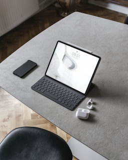 A tablet with a keyboard is placed on a minimalist, gray tabletop. Accompanying it are a smartphone, a pair of wireless earbuds, and their charging case. The surface is smooth and sleek, complemented by a black chair partially visible at the bottom of the image.