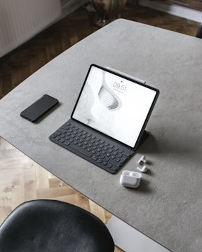 A tablet with a keyboard is placed on a minimalist, gray tabletop. Accompanying it are a smartphone, a pair of wireless earbuds, and their charging case. The surface is smooth and sleek, complemented by a black chair partially visible at the bottom of the image.
