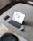A tablet with a keyboard is placed on a minimalist, gray tabletop. Accompanying it are a smartphone, a pair of wireless earbuds, and their charging case. The surface is smooth and sleek, complemented by a black chair partially visible at the bottom of the image.
