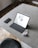 A tablet with a keyboard is placed on a minimalist, gray tabletop. Accompanying it are a smartphone, a pair of wireless earbuds, and their charging case. The surface is smooth and sleek, complemented by a black chair partially visible at the bottom of the image.