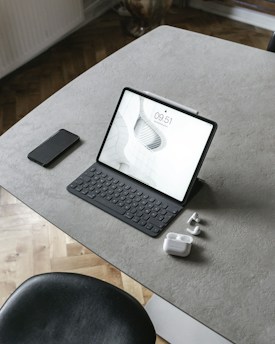A tablet with a keyboard is placed on a minimalist, gray tabletop. Accompanying it are a smartphone, a pair of wireless earbuds, and their charging case. The surface is smooth and sleek, complemented by a black chair partially visible at the bottom of the image.