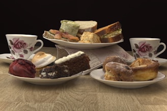 A variety of pastries and sandwiches are arranged on plates, with two floral teacups placed on either side. The pastries include a red velvet treat, a piece of chocolate cake, a cream-topped brownie, and assorted baked goods like scones. The sandwiches appear to be made with different types of bread and fillings, and everything is displayed on a wooden table with a dark background.