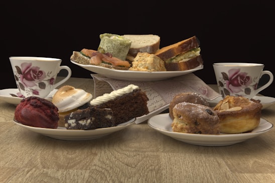 A variety of pastries and sandwiches are arranged on plates, with two floral teacups placed on either side. The pastries include a red velvet treat, a piece of chocolate cake, a cream-topped brownie, and assorted baked goods like scones. The sandwiches appear to be made with different types of bread and fillings, and everything is displayed on a wooden table with a dark background.