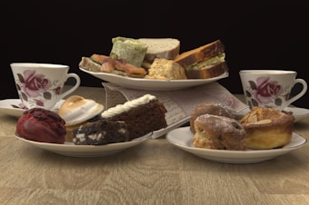 A variety of pastries and sandwiches are arranged on plates, with two floral teacups placed on either side. The pastries include a red velvet treat, a piece of chocolate cake, a cream-topped brownie, and assorted baked goods like scones. The sandwiches appear to be made with different types of bread and fillings, and everything is displayed on a wooden table with a dark background.