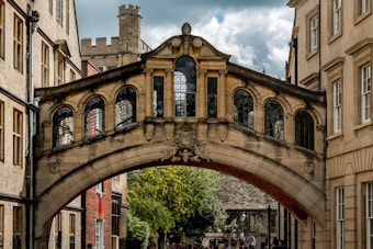 A historic stone bridge connects two buildings with ornate architectural details and arched windows. The bridge is elevated over a pedestrian passage with a group of people walking below on a street flanked by traditional building facades with many windows.