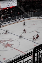 A professional ice hockey match captured from a high vantage point, featuring players in red and white uniforms moving across the rink. The scene includes a large digital screen displaying a live feed of the game and a sparsely filled audience watching from the stands.