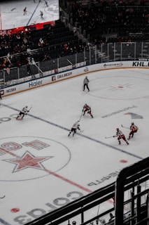 A professional ice hockey match captured from a high vantage point, featuring players in red and white uniforms moving across the rink. The scene includes a large digital screen displaying a live feed of the game and a sparsely filled audience watching from the stands.