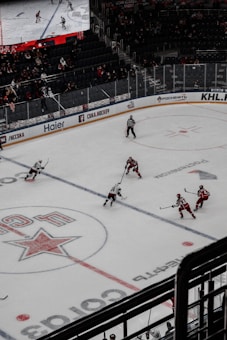 A professional ice hockey match captured from a high vantage point, featuring players in red and white uniforms moving across the rink. The scene includes a large digital screen displaying a live feed of the game and a sparsely filled audience watching from the stands.