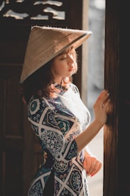 A woman wearing a patterned dress is bathed in warm sunlight while standing near a wooden door. She wears a traditional conical hat and holds her hand against the doorframe.
