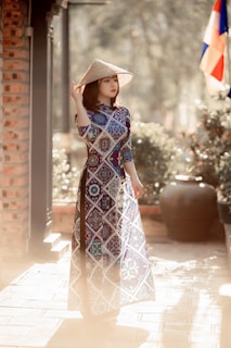 A person stands outdoors wearing a traditional patterned long dress, holding a conical hat. The scene is enriched with soft lighting, and a flag is visible in the background. Surrounding the figure are elements of nature and architectural details, lending a harmonious blend of culture and environment.