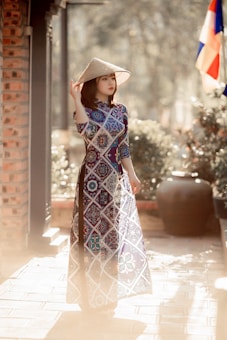 A person stands outdoors wearing a traditional patterned long dress, holding a conical hat. The scene is enriched with soft lighting, and a flag is visible in the background. Surrounding the figure are elements of nature and architectural details, lending a harmonious blend of culture and environment.