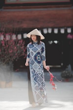 A woman wearing a traditional Vietnamese áo dài dress featuring blue and white patterns stands elegantly, with a conical hat on her head. In her hand, she holds a festive decoration with red tassels. The background includes a blurred view of a building and blossoming trees, suggesting a culturally rich setting.