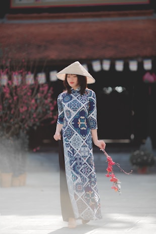 A woman wearing a traditional Vietnamese áo dài dress featuring blue and white patterns stands elegantly, with a conical hat on her head. In her hand, she holds a festive decoration with red tassels. The background includes a blurred view of a building and blossoming trees, suggesting a culturally rich setting.