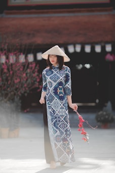 A woman wearing a traditional Vietnamese áo dài dress featuring blue and white patterns stands elegantly, with a conical hat on her head. In her hand, she holds a festive decoration with red tassels. The background includes a blurred view of a building and blossoming trees, suggesting a culturally rich setting.