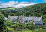 A picturesque rural scene featuring traditional cottages with slate roofs situated in a lush green landscape. A viaduct spans the background, set against rolling hills and dense woodlands. The foreground includes well-tended gardens with various plants and a wooden fence.