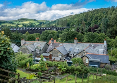 A picturesque rural scene featuring traditional cottages with slate roofs situated in a lush green landscape. A viaduct spans the background, set against rolling hills and dense woodlands. The foreground includes well-tended gardens with various plants and a wooden fence.