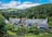A picturesque rural scene featuring traditional cottages with slate roofs situated in a lush green landscape. A viaduct spans the background, set against rolling hills and dense woodlands. The foreground includes well-tended gardens with various plants and a wooden fence.