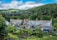 A picturesque rural scene featuring traditional cottages with slate roofs situated in a lush green landscape. A viaduct spans the background, set against rolling hills and dense woodlands. The foreground includes well-tended gardens with various plants and a wooden fence.