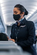 A flight attendant wearing a face mask and a navy uniform, standing in the aisle of an airplane, engaged in assisting passengers. Her hair is neatly tied back, and she wears a name tag and a scarf. The airplane cabin has a modern design with overhead lighting.