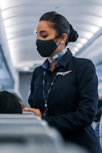 A flight attendant wearing a face mask and a navy uniform, standing in the aisle of an airplane, engaged in assisting passengers. Her hair is neatly tied back, and she wears a name tag and a scarf. The airplane cabin has a modern design with overhead lighting.