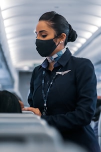 A flight attendant wearing a face mask and a navy uniform, standing in the aisle of an airplane, engaged in assisting passengers. Her hair is neatly tied back, and she wears a name tag and a scarf. The airplane cabin has a modern design with overhead lighting.