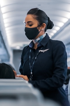 A flight attendant wearing a face mask and a navy uniform, standing in the aisle of an airplane, engaged in assisting passengers. Her hair is neatly tied back, and she wears a name tag and a scarf. The airplane cabin has a modern design with overhead lighting.