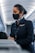 A flight attendant wearing a face mask and a navy uniform, standing in the aisle of an airplane, engaged in assisting passengers. Her hair is neatly tied back, and she wears a name tag and a scarf. The airplane cabin has a modern design with overhead lighting.