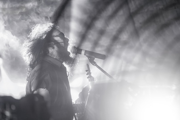 A black and white image of a musician passionately singing into a microphone. The scene is dramatic, with swirling smoke and intense lighting effects surrounding the artist, who has long hair and an expressive facial expression.