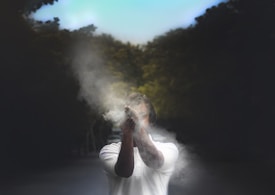 A person stands in the center of a dark, tree-lined background, wearing a white shirt and clapping hands together, resulting in a cloud of dust or smoke enveloping their hands and face.