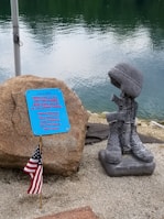 A memorial display features a stone with a plaque dedicated to men and women who served in the armed forces. Beside the stone, a sculpture consisting of military boots, a rifle, and a helmet symbolizes a battlefield cross. An American flag is planted next to it. The background shows a body of water with gentle ripples.