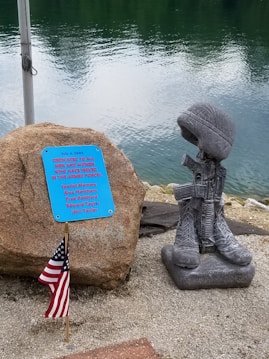 A memorial display features a stone with a plaque dedicated to men and women who served in the armed forces. Beside the stone, a sculpture consisting of military boots, a rifle, and a helmet symbolizes a battlefield cross. An American flag is planted next to it. The background shows a body of water with gentle ripples.