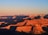The expansive view of the Grand Canyon highlights its layered red, orange, and brown rock formations, bathed in the warm glow of sunset. Shadows accentuate the ridges and valleys, creating a striking contrast against the clear blue sky.