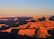 The expansive view of the Grand Canyon highlights its layered red, orange, and brown rock formations, bathed in the warm glow of sunset. Shadows accentuate the ridges and valleys, creating a striking contrast against the clear blue sky.