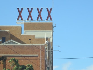 A large red sign consisting of four Xs is mounted on the roof of an industrial-looking brick building. The building features geometric architectural details, such as a triangular window and rectangular forms. In the foreground, there are green trees and power lines, with a clear blue sky in the background.