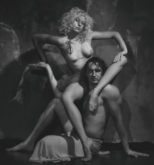 A black and white photograph features a man and a woman in an artistic pose. The woman has curly hair and is wearing a bikini top, sitting on the man's shoulder with her legs elegantly posed. The man is shirtless, and both display an expressive posture against a textured wall.