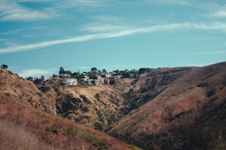 A hillside with dry vegetation is contrasted by a cluster of houses perched at the top. The terrain is rugged and shows signs of erosion, with the sky above being clear and blue, scattered with light clouds. The houses are surrounded by trees, adding a touch of greenery to the scene.