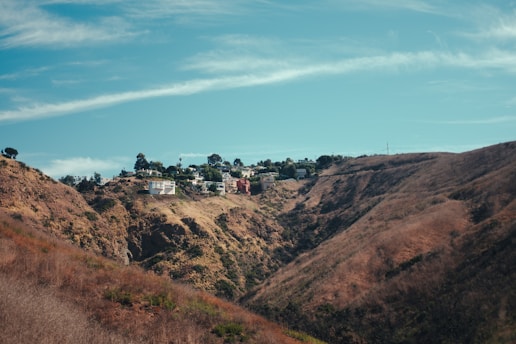 A hillside with dry vegetation is contrasted by a cluster of houses perched at the top. The terrain is rugged and shows signs of erosion, with the sky above being clear and blue, scattered with light clouds. The houses are surrounded by trees, adding a touch of greenery to the scene.