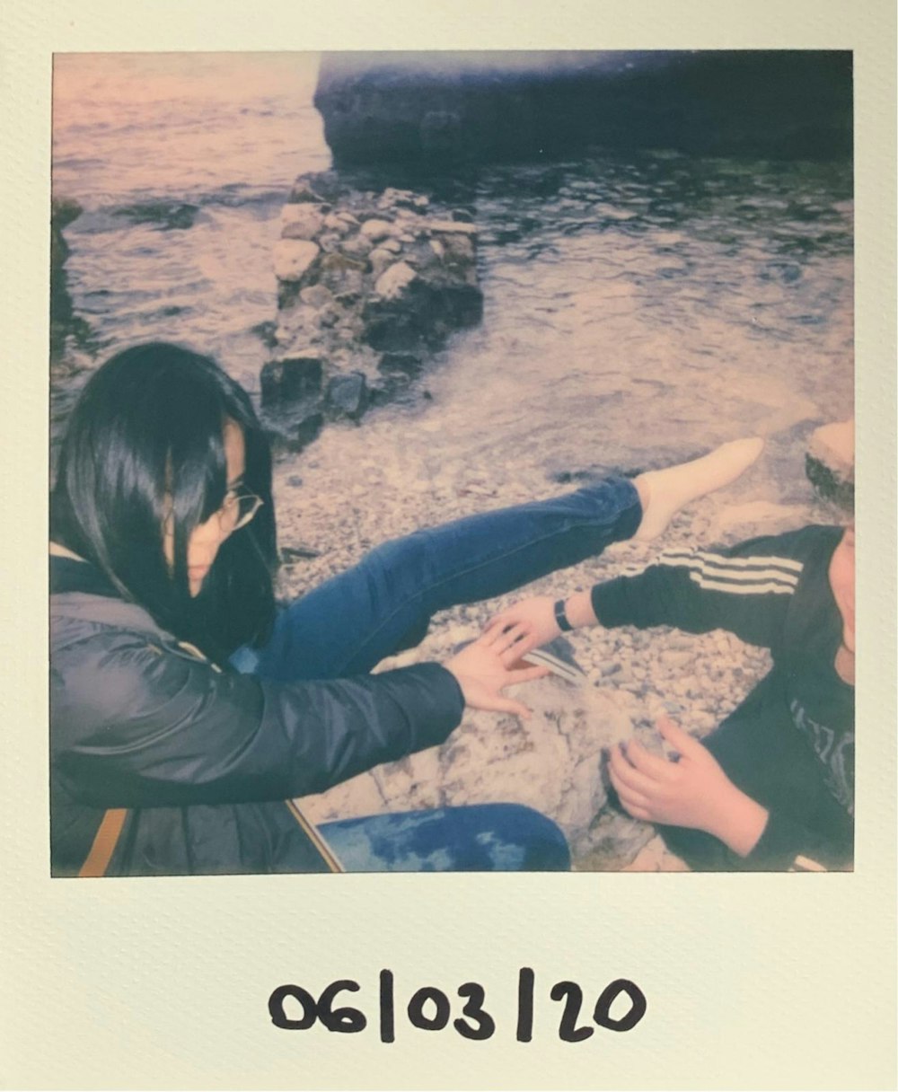 woman in black jacket and blue denim jeans sitting on rock near body of water during