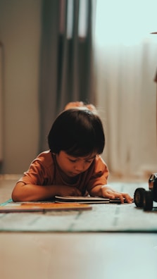 A young child is lying on the floor with focus and attention directed toward a book in front of them. The atmosphere is calm with natural light coming through a window, softly illuminating the scene. Nearby, a toy car is also visible, adding a playful element to the setting.