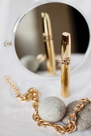 A gold-colored cylindrical object, possibly a cosmetic product, stands upright in front of a circular mirror, reflecting its image. Nearby, a gold chain is elegantly placed beside two smooth gray stones on a white surface.