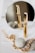 A gold-colored cylindrical object, possibly a cosmetic product, stands upright in front of a circular mirror, reflecting its image. Nearby, a gold chain is elegantly placed beside two smooth gray stones on a white surface.